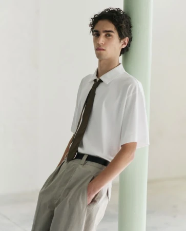 Model leaning against a pale green column wearing a white short-sleeve collared shirt, dark tie, and light trousers.