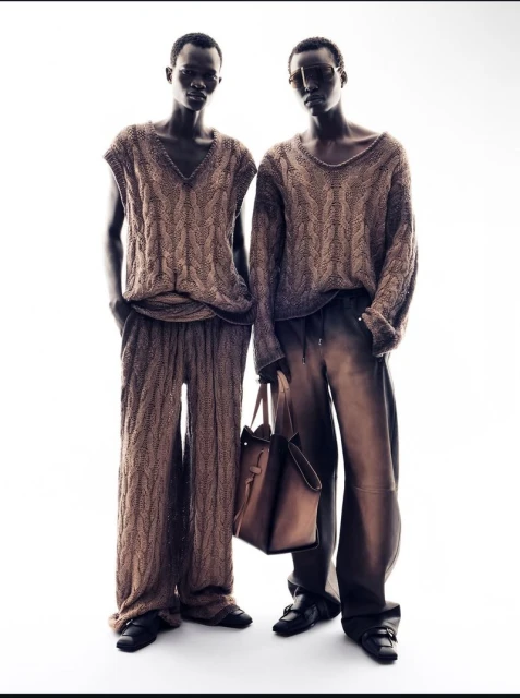 Two dark-skinned male fashion models in matching knit outfits, standing in front of a bright white background, accessorized with a handbag.