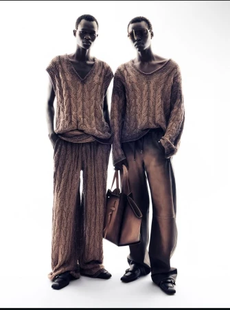 Two dark-skinned male fashion models in matching knit outfits, standing in front of a bright white background, accessorized with a handbag.