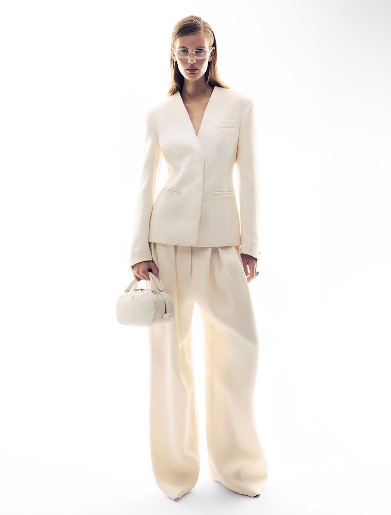 Fashion shot of a woman in a light-cream coloured suit holding a handbag, standing in a bright photo studio with a white background.