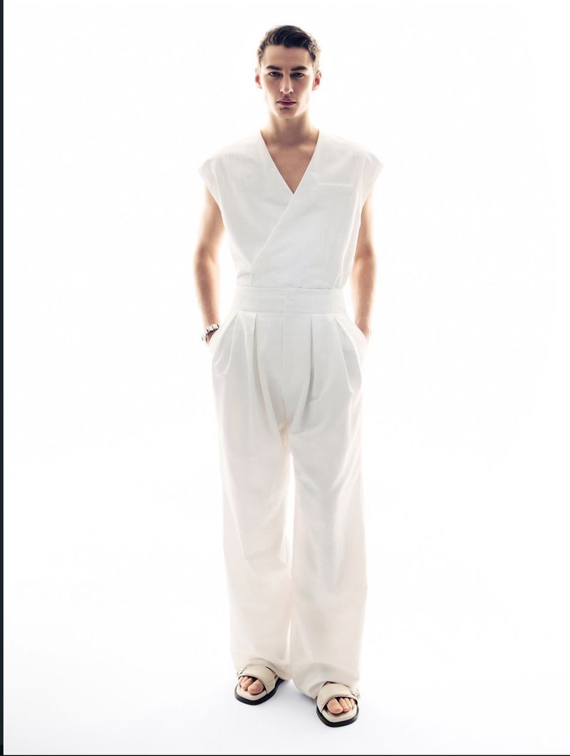 Full-length shot of a young adult male model in a white wrap top and wide-leg pants, posing with hands in pockets against a bright white background.