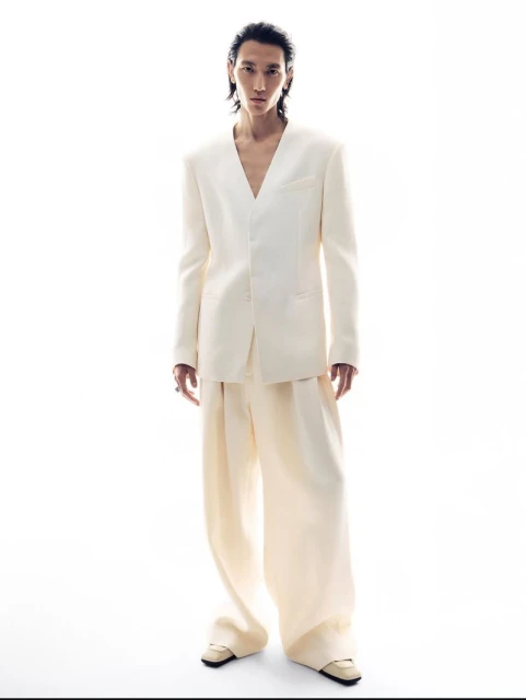 An adult male poses in a fashion editorial shot, wearing a light suit and wide-legged pants against a bright white backdrop.