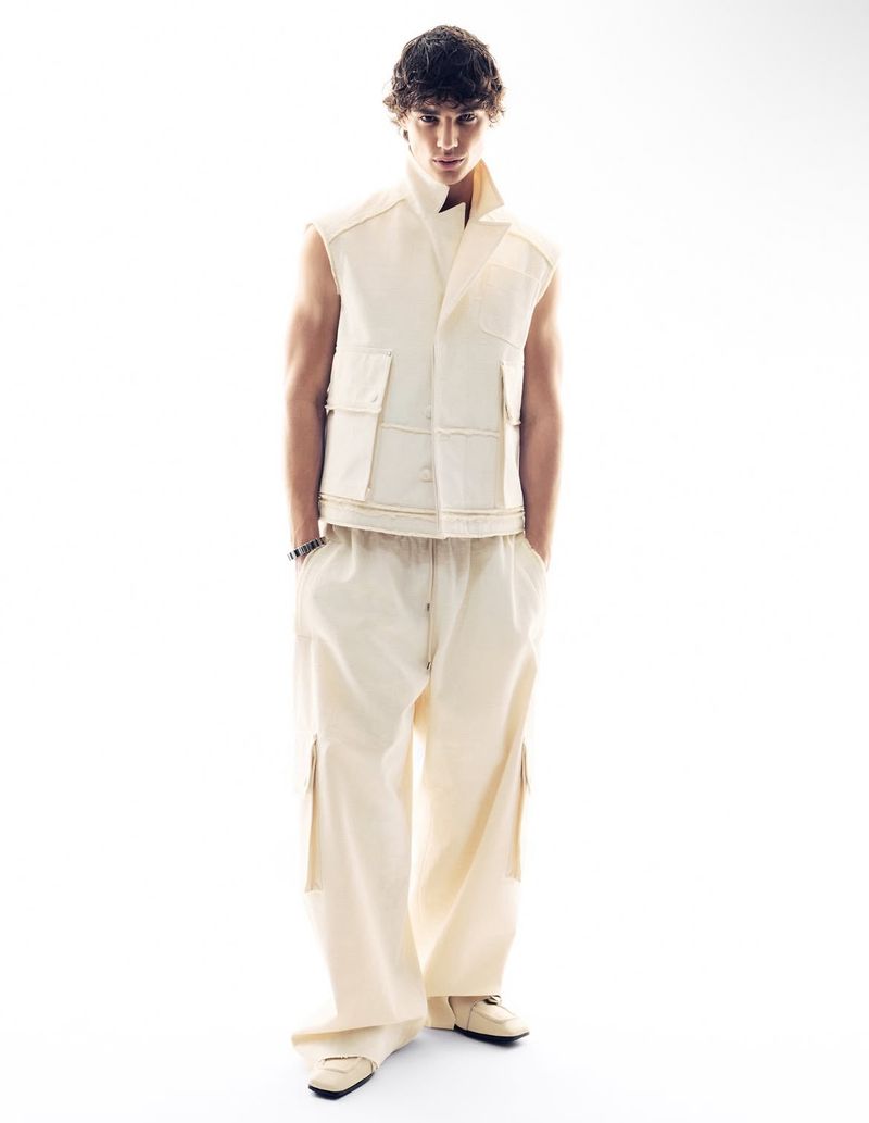 Full-length shot of a young adult male fashion model in an off-white vest and baggy pants against a bright white backdrop in a photo studio.