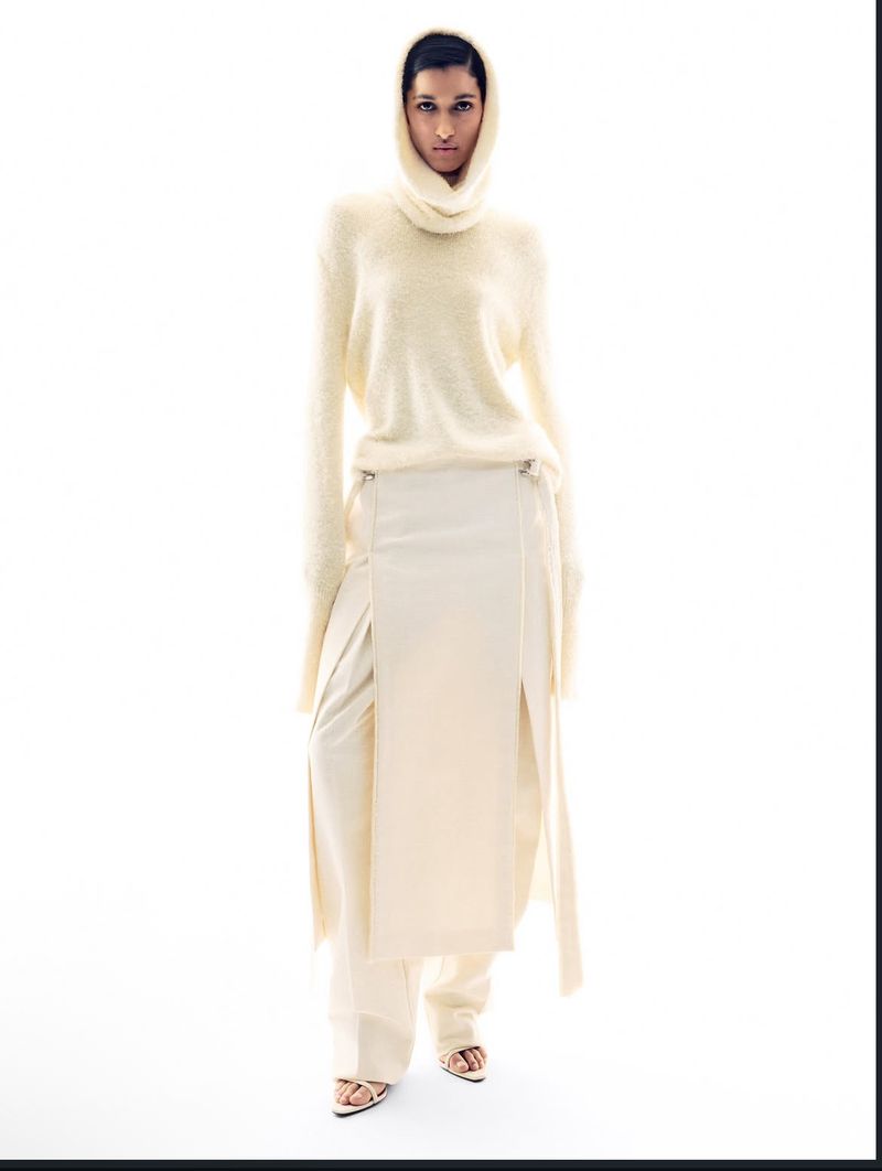 Fashion shot of a young adult woman with fair skin, wearing a beige sweater with high neck, long skirt, and sandals on a white background.