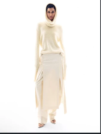 Fashion shot of a young adult woman with fair skin, wearing a beige sweater with high neck, long skirt, and sandals on a white background.