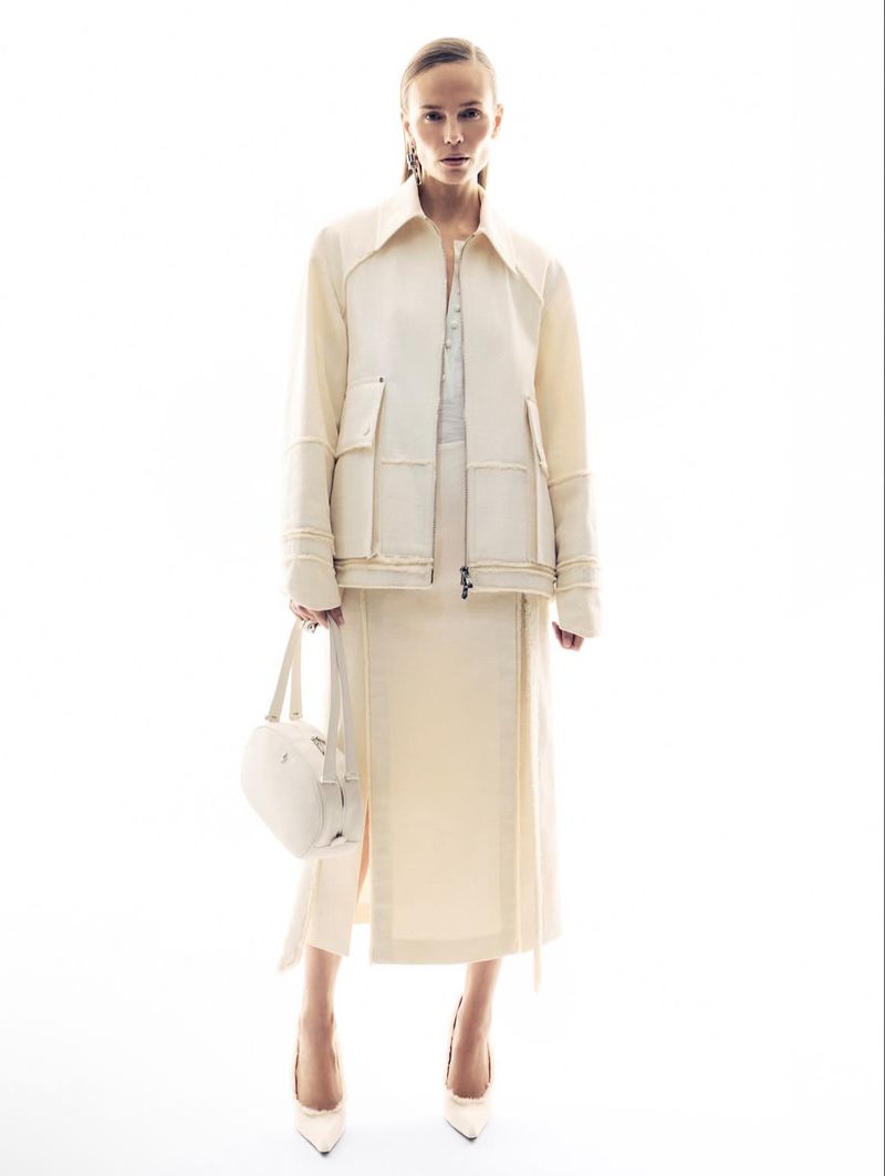 Model in cream jacket and matching midi skirt holding a white handbag against a plain white background