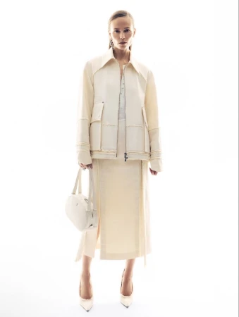 Model in cream jacket and matching midi skirt holding a white handbag against a plain white background