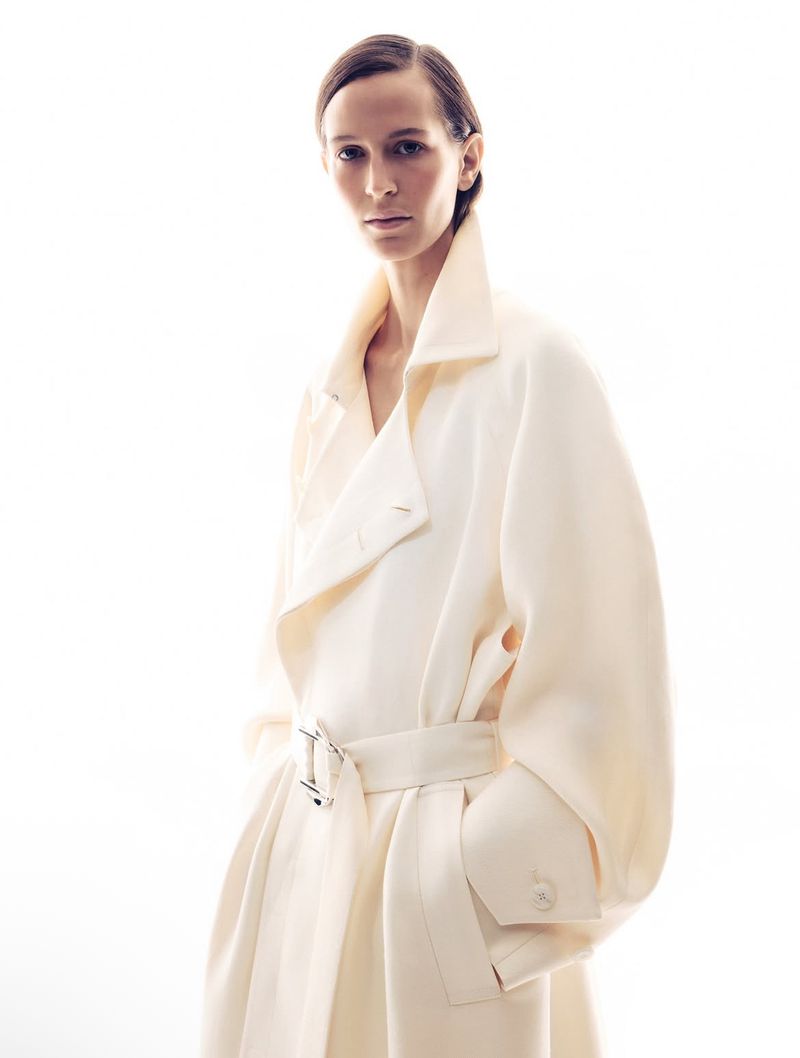 Model wearing a cream-colored belted coat with a high collar against a bright white background.