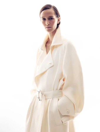 Model wearing a cream-colored belted coat with a high collar against a bright white background.