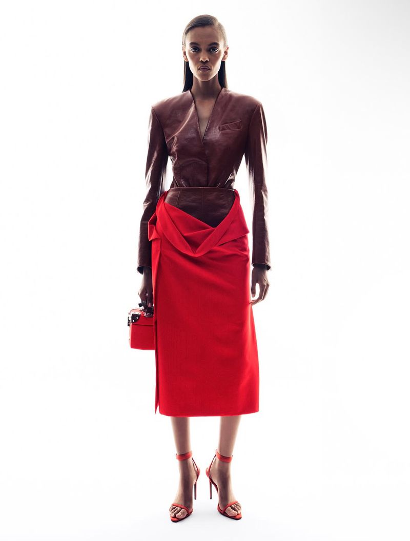 A brown-skinned woman poses in a studio wearing a brown leather top, red skirt, and red high heels while holding a small red purse.
