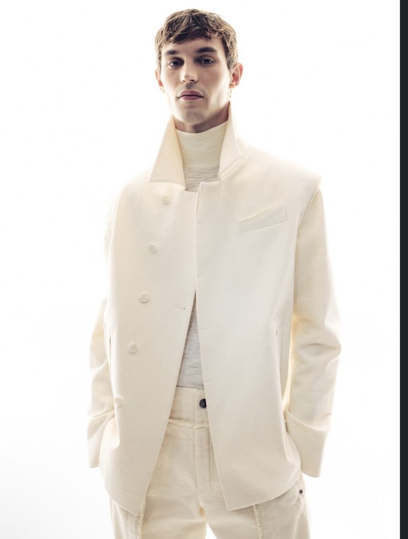 A fashionable young adult male poses in a white jacket and matching pants against a bright, high-key background in a studio setting.