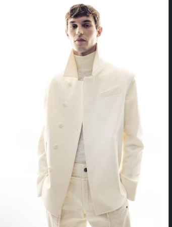 A fashionable young adult male poses in a white jacket and matching pants against a bright, high-key background in a studio setting.