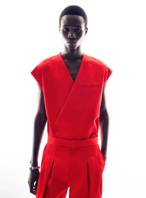 Studio shot of an adult male fashion model in a bright red sleeveless top and matching pants against a white background, minimalist style.