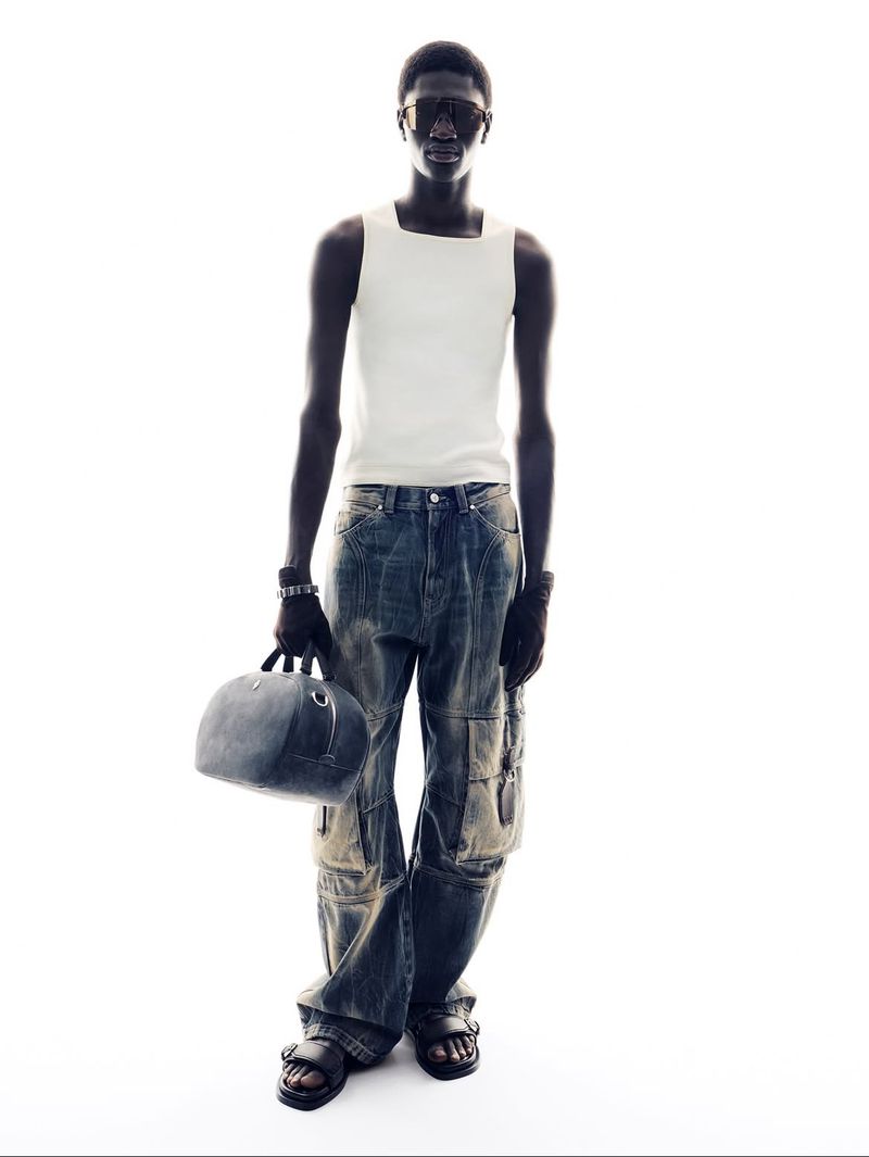 Full-length shot of a Black male model wearing a white tank top, denim cargo pants, and sunglasses, holding a round bag against a bright white background.