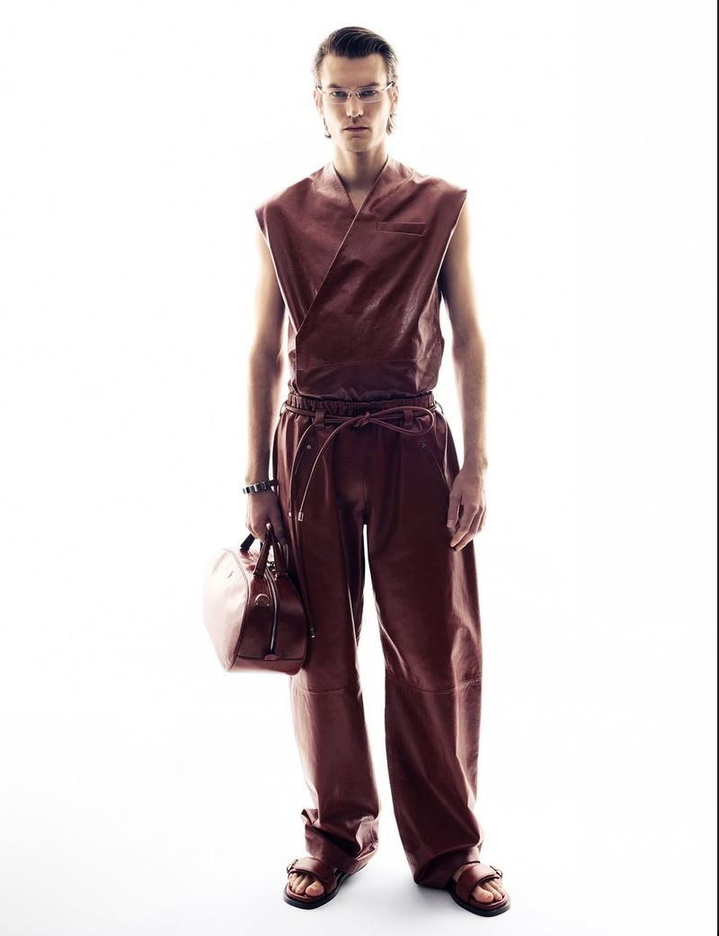 Full-length studio shot of a man in brown sleeveless top and pants holding a matching bag, eyeglasses, and sandals against white backdrop.