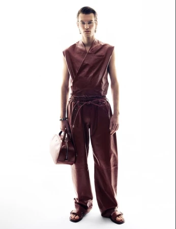 Full-length studio shot of a man in brown sleeveless top and pants holding a matching bag, eyeglasses, and sandals against white backdrop.