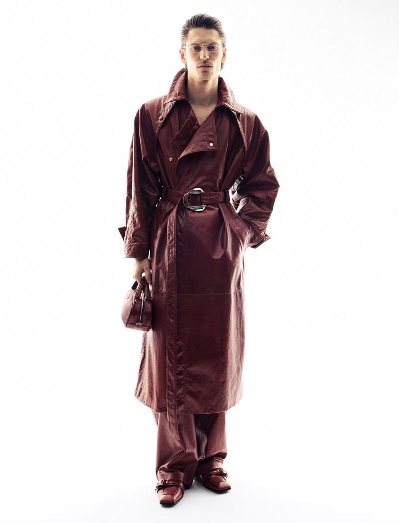 Full-length studio shot of a person modeling a reddish-brown, belted trench coat with matching pants, shoes, and bag against white backdrop.