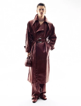 Full-length studio shot of a person modeling a reddish-brown, belted trench coat with matching pants, shoes, and bag against white backdrop.