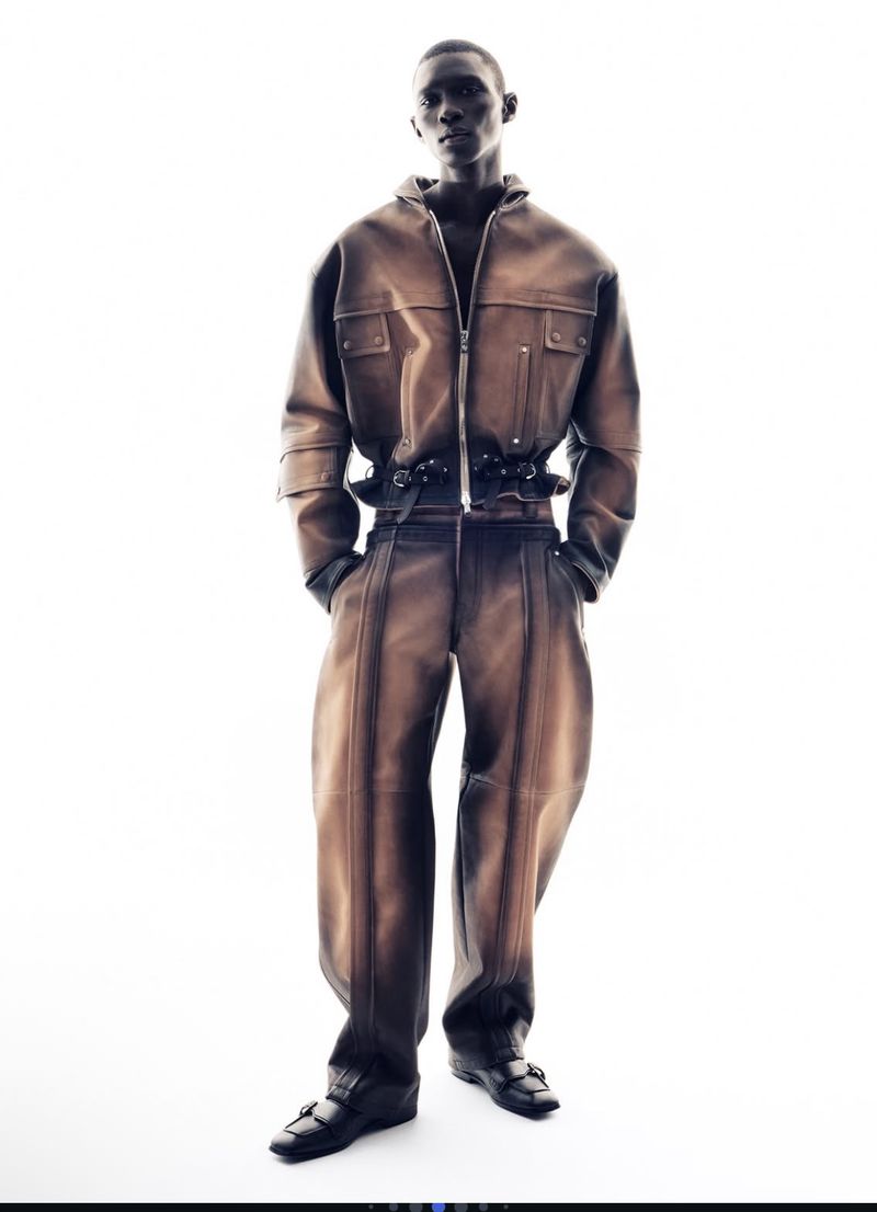 Full-length studio shot of an adult male fashion model in a tan leather jumpsuit, posing against a bright white background with hands in pockets.