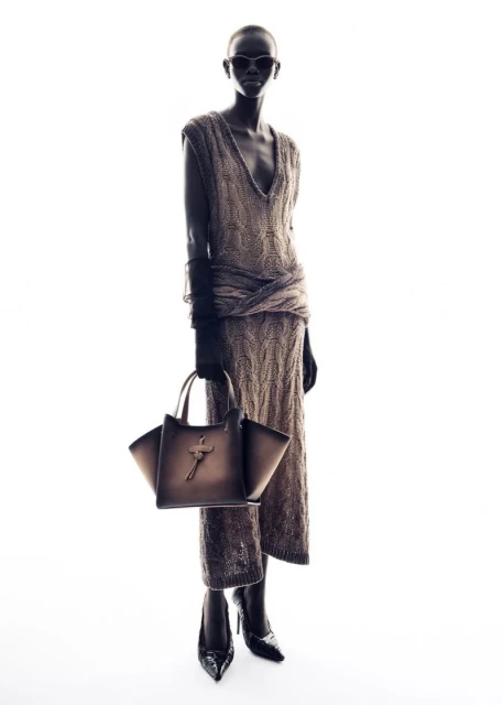 Full-length fashion portrait of an adult female model wearing a knitted sweater dress, sunglasses, heels, and carrying a handbag.
