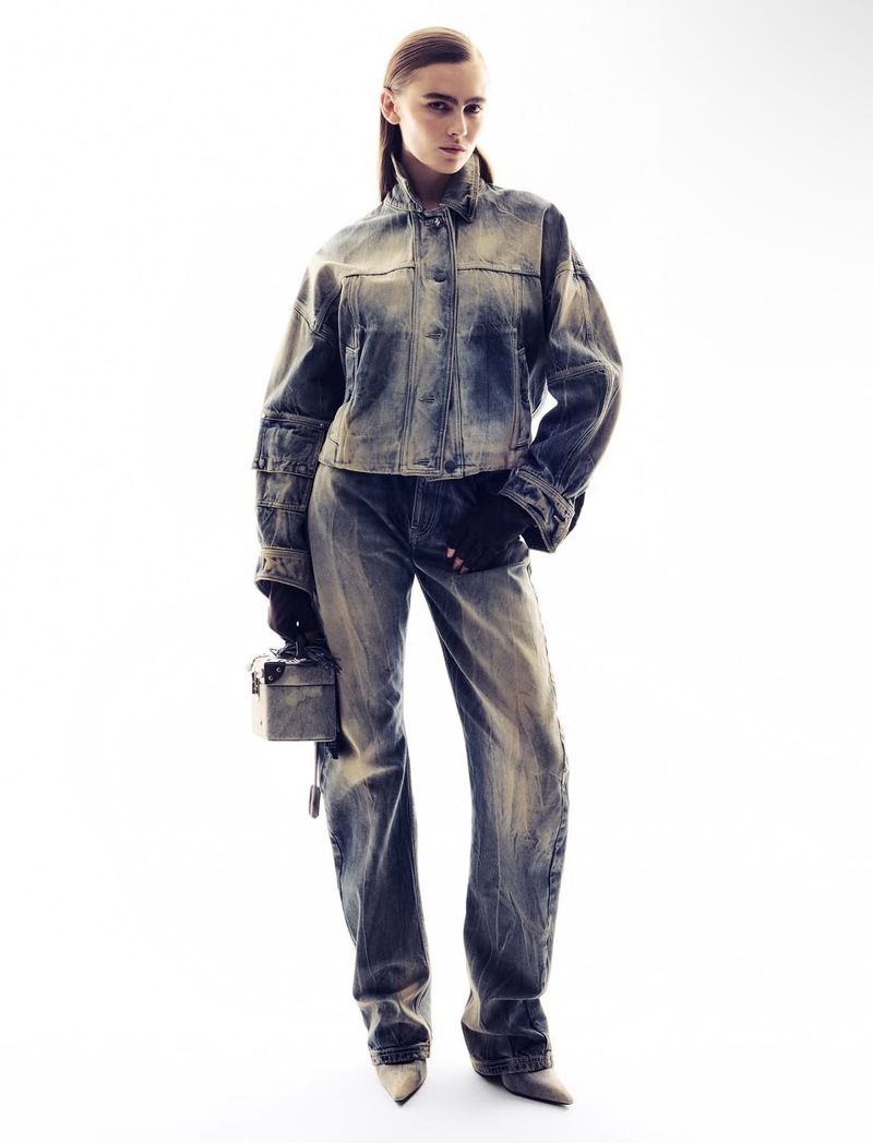 A woman posing in an acid-wash denim jacket and jeans, with beige heels, in a brightly lit studio setting.