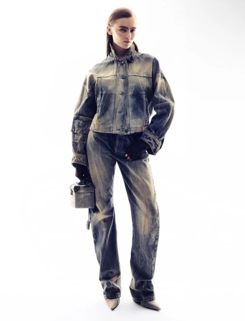 A woman posing in an acid-wash denim jacket and jeans, with beige heels, in a brightly lit studio setting.