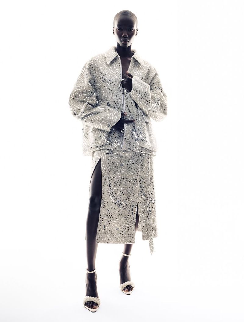 Model standing against a pure white background wearing a sparkling patterned metallic silver jacket and matching skirt.