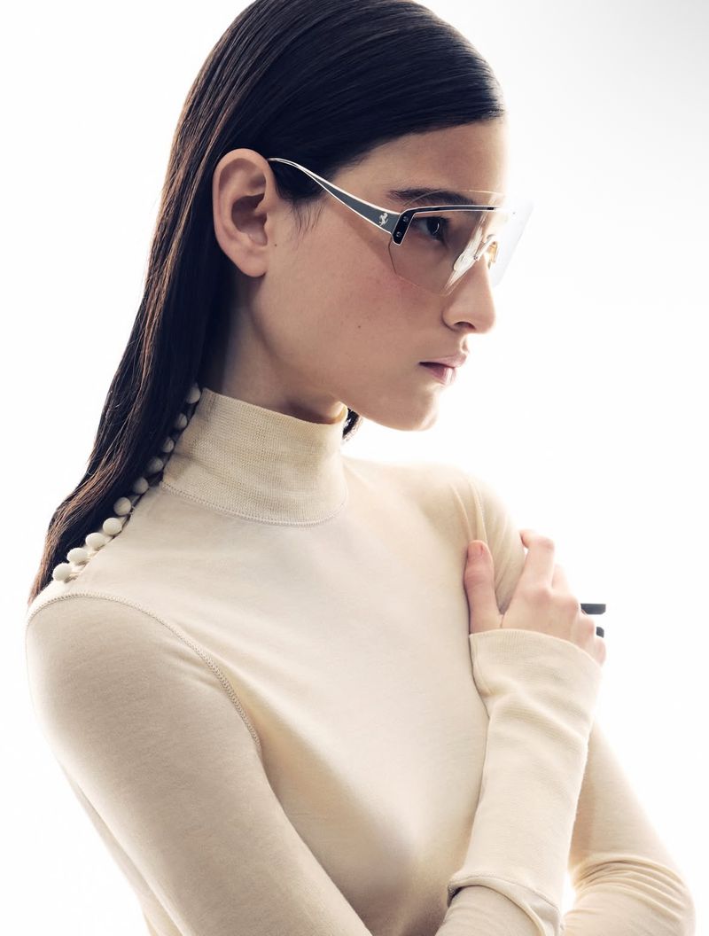 Close-up studio shot of a female model wearing stylish eyewear and a beige turtleneck sweater with button details on the sleeve.