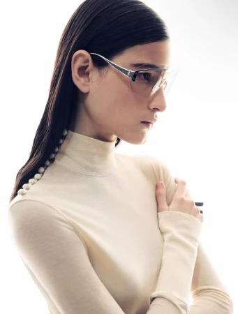 Close-up studio shot of a female model wearing stylish eyewear and a beige turtleneck sweater with button details on the sleeve.