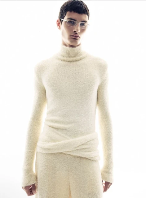 Close-up of a slender man in a cream-colored, textured turtleneck and pants posing against a bright white background, wearing wire-frame glasses.