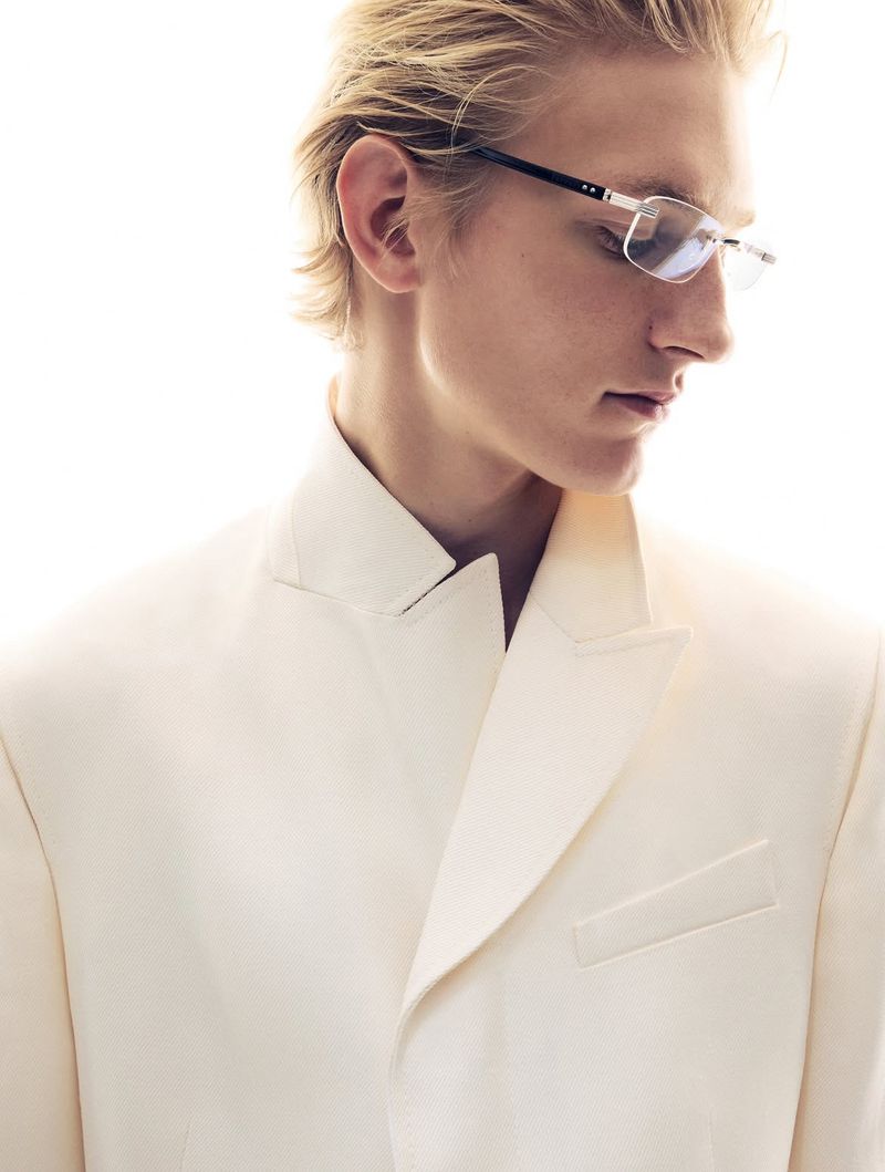 Elegant man in a white suit and glasses, posing in a studio environment with bright lighting, creating a sophisticated fashion portrait.