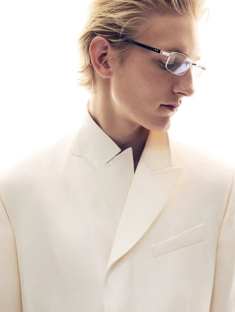 Elegant man in a white suit and glasses, posing in a studio environment with bright lighting, creating a sophisticated fashion portrait.