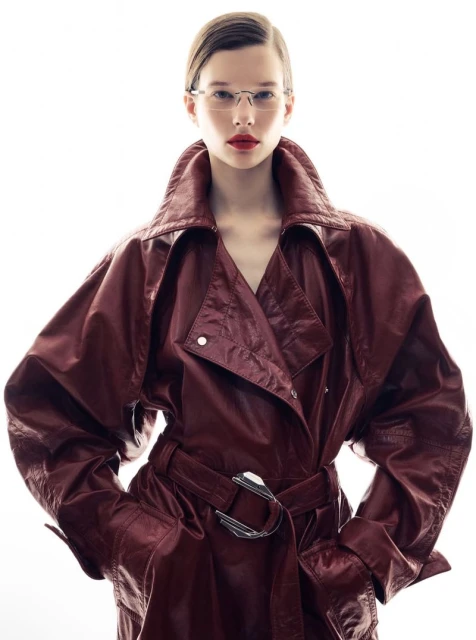 Stylish woman with glasses and a red leather trench coat posing in a bright, white studio. Fashion photography showcasing the texture of the coat.