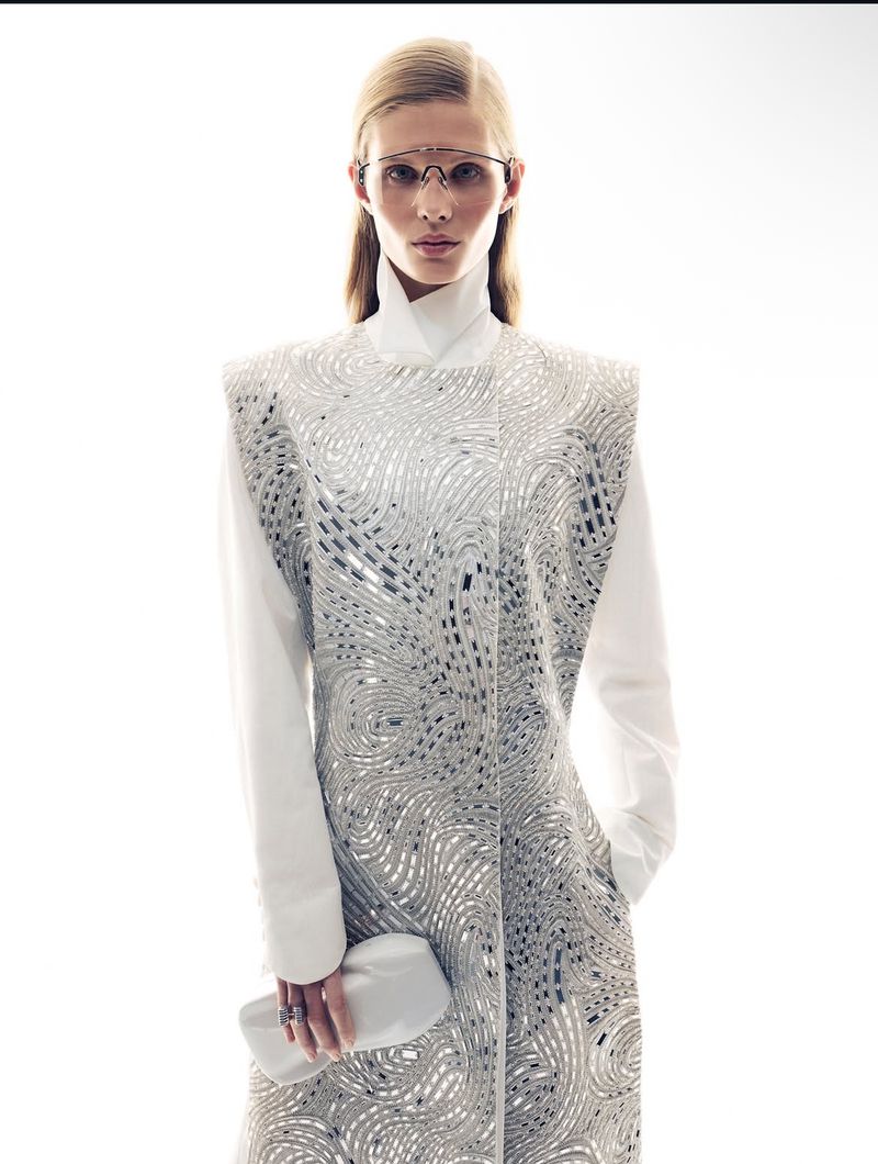Blonde woman in white dress with silver detailing, posing in a studio setting with a white clutch and futuristic glasses.