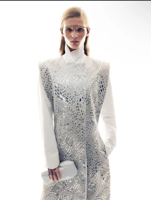 Blonde woman in white dress with silver detailing, posing in a studio setting with a white clutch and futuristic glasses.