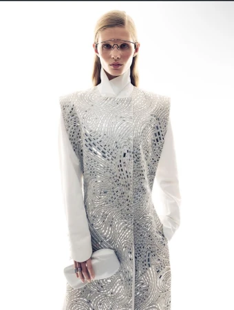 Model wearing a patterned sleeveless coat and white shirt, holding a white clutch against a plain white background.