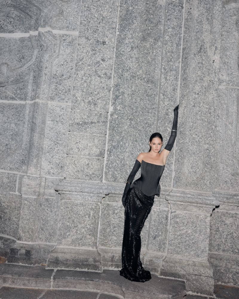An adult female in a black gown and gloves poses leaning against a grey stone wall; the ensemble features an off-shoulder corset.