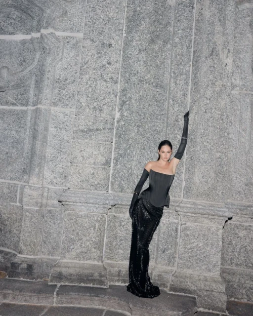 An adult female in a black gown and gloves poses leaning against a grey stone wall; the ensemble features an off-shoulder corset.