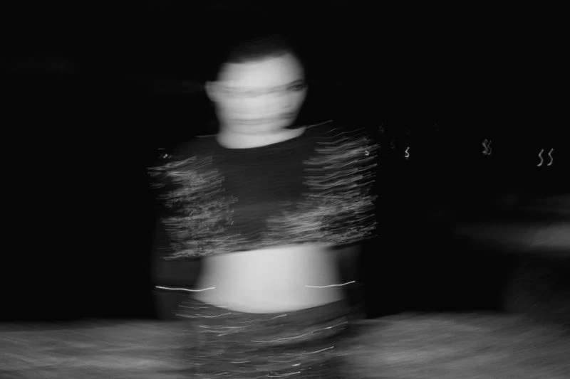 Blurred black and white image of an adult female, wearing a top, with a dark background, and motion blur.
