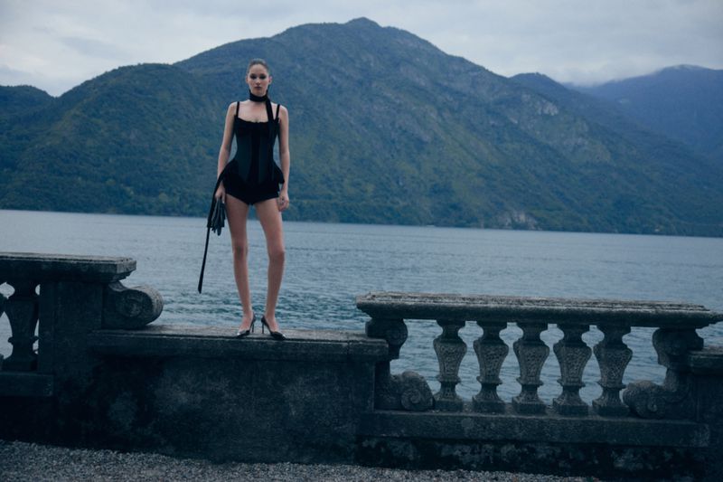 A woman in black lingerie stands on a stone ledge, overlooking a lake and mountains, creating a dramatic fashion portrait in nature.