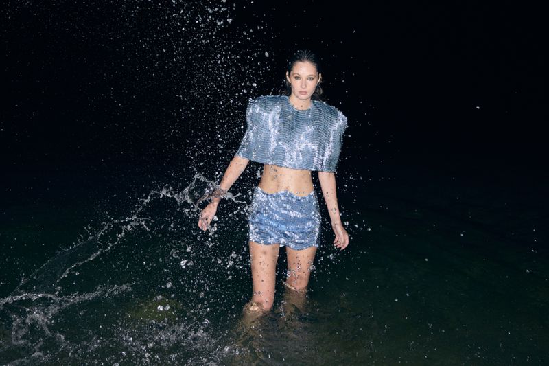 An adult woman poses waist-deep in dark water, wearing a shiny silver crop top and shorts. Splashes of water surround her in the dark.