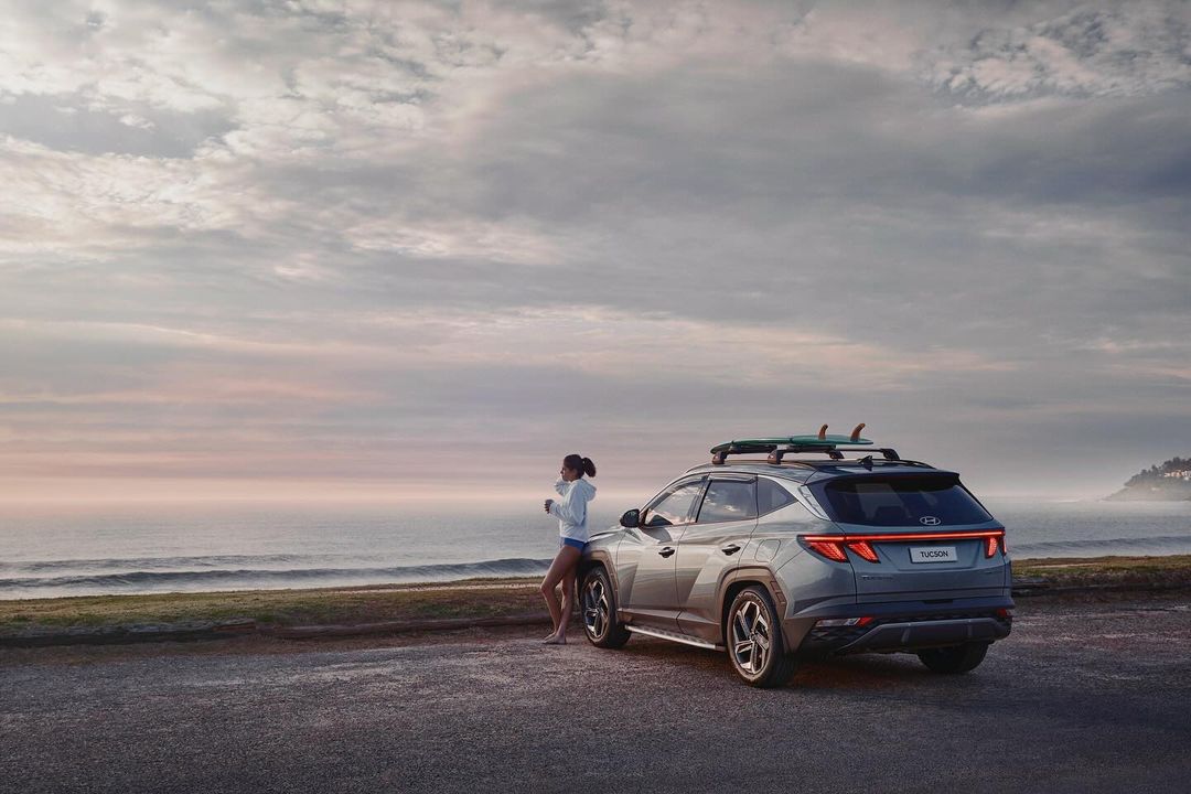 Grey Hyundai Tucson SUV parked near a beach at sunset with a woman leaning on the hood with a surfboard on the roof rack.