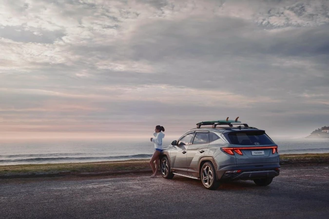 Grey Hyundai Tucson SUV parked near a beach at sunset with a woman leaning on the hood with a surfboard on the roof rack.