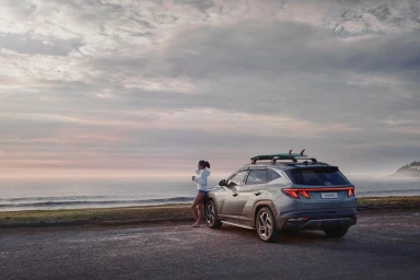 A woman leaning against a grey Hyundai SUV parked near a coastal beach under an overcast sunset sky