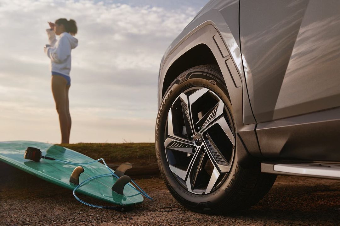 Hyundai Tucson SUV parked on beach with woman surfer looking out at the ocean and surfboard laying on the sand in the foreground.