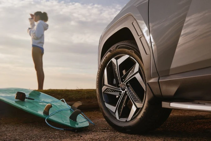 Hyundai Tucson SUV parked on beach with woman surfer looking out at the ocean and surfboard laying on the sand in the foreground.