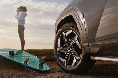 Close-up of a modern grey SUV wheel with a surf board lying on the ground and a person standing in the background.