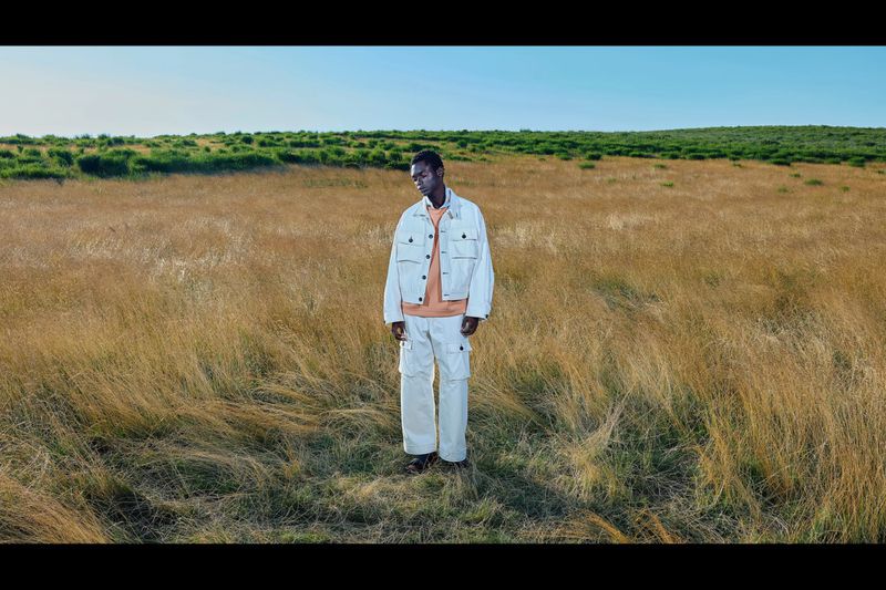 A lone Black male model stands in a golden field wearing a white jacket and pants, under a clear sky with a grassy hill in the distance.