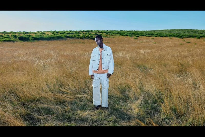 A lone Black male model stands in a golden field wearing a white jacket and pants, under a clear sky with a grassy hill in the distance.