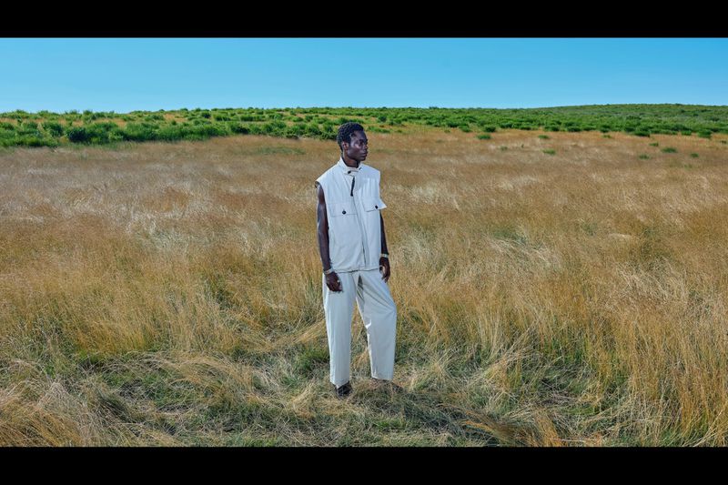 A solitary figure stands in a windswept field under a clear sky, wearing a light-colored vest and trousers, evoking a sense of serene isolation.